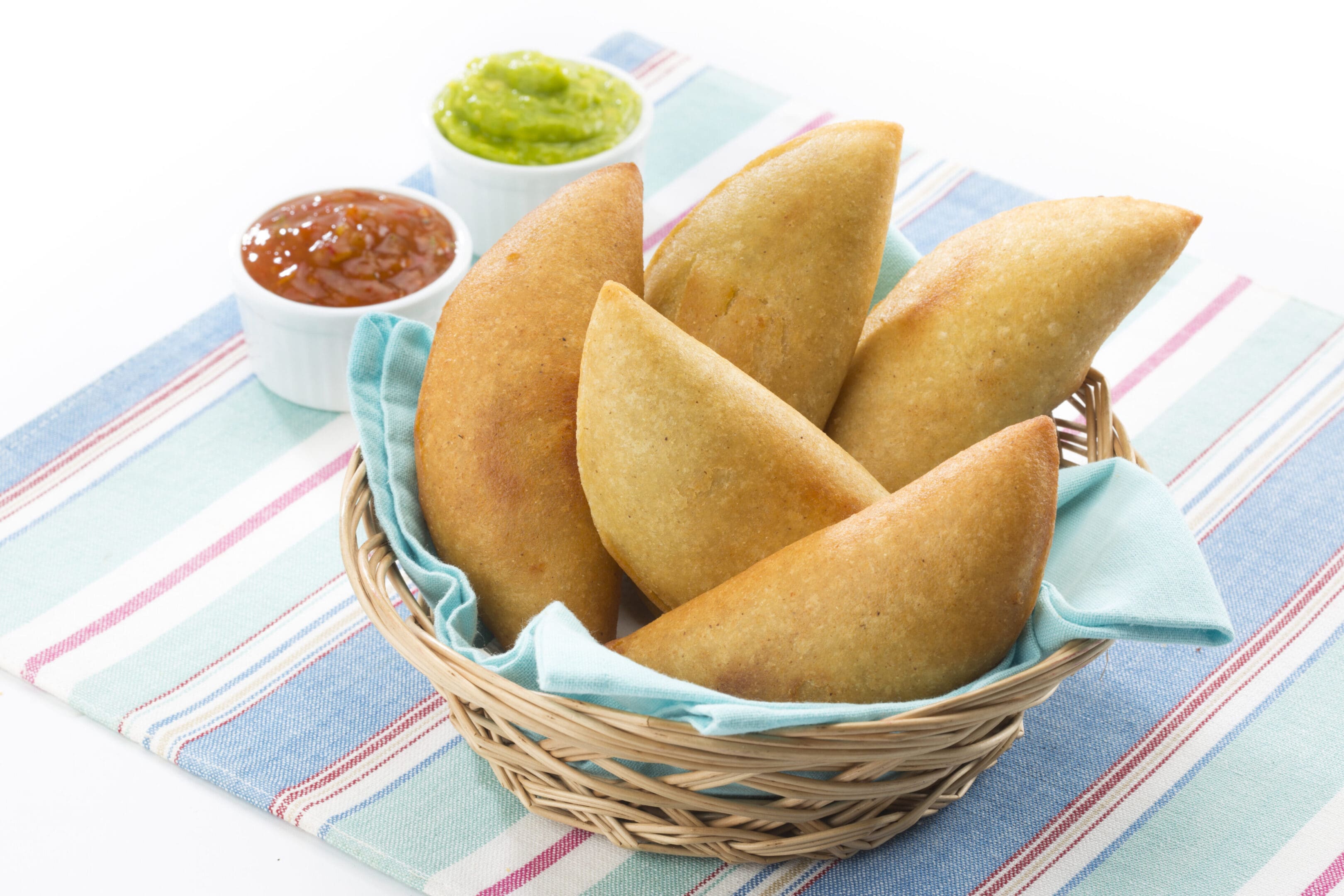 Basket of golden empanadas with dipping sauces on a striped cloth.