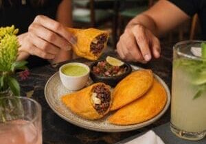 Hands reaching for plated golden empanadas with shredded beef, plantain and cheese - Illustrative Image